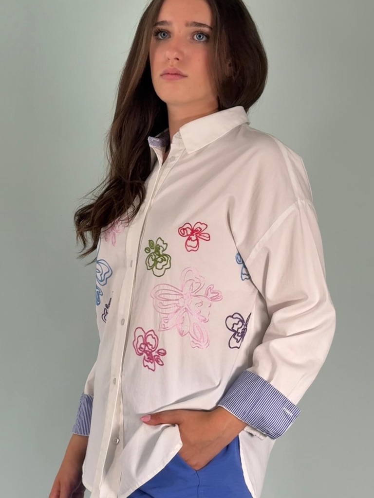 Camicia ricamata, con il dettaglio del polsino con risvolto a righe bianche e blu. Allacciata sul davanti con bottoni a forma di fiore.