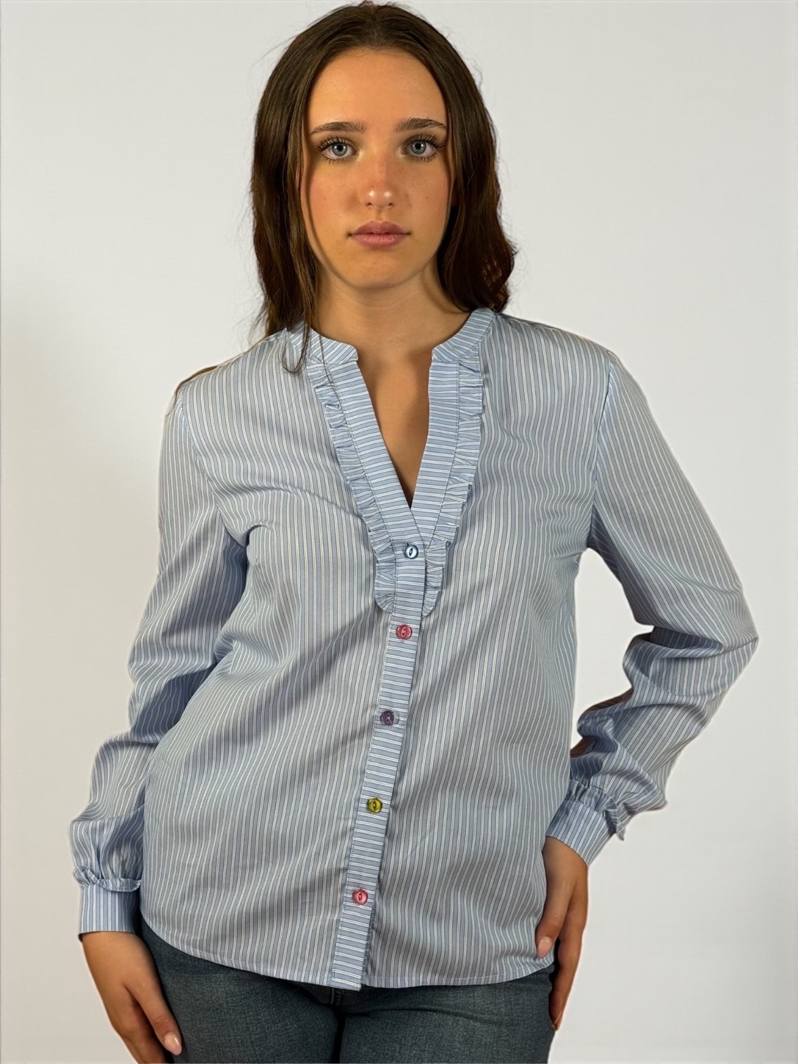 Camicia a righe celeste, allacciata sul davanti con bottoni di diversi colori. Scollatura a V manica lunga.